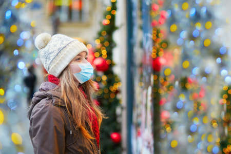 Girl wearing face mask on a Parisian street or at Christmas market looking at shop windows decorated for Christmas. Seasonal holidays during pandemic and coronavirus outbreakの写真素材