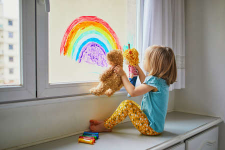 Toddler girl drawing rainbow on window glass as sign of hope and showing it to her teddy and doll. Creative games for kids staying at home during lockdown. Self isolation and coronavirus quarantineの写真素材