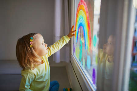 Adorable toddler girl drawing rainbow on window glass as sign of hope. Creative games for kids staying at home during lockdown. Self isolation and coronavirus quarantine conceptの写真素材