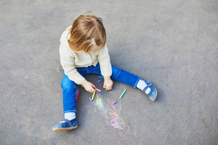 Adorable toddler girl drawing with colorful chalks on asphalt. Outdoor activity and creative games for small kidsの写真素材