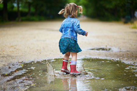 Child wearing red rain boots and jumping in puddle on a summer day. Adorable toddler girl having fun with water and mud in park on a rainy day. Outdoor activities for kidsの写真素材