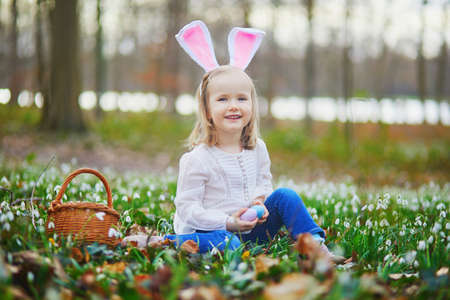 Girl wearing bunny ears playing egg hunt on Easter. Toddler sitting on the grass with many snowdrop...