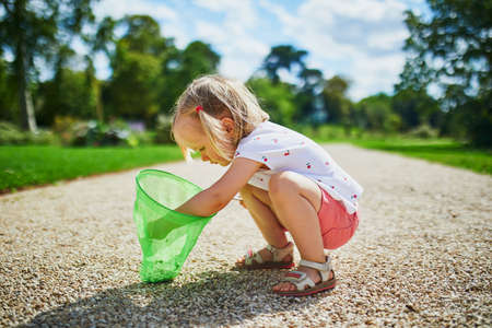 Adorable toddler girl playing with butterfy net outdoors on a summer day. Child gathering pebbles in scoop-net. Outdoor summer activities for kids.の写真素材