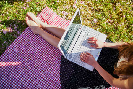 Beautiful young girl working on her laptop in park during cherry blossom season. Young woman in famous Park of Sceaux near Paris, France. Freelance, distance learning or remote work conceptの写真素材