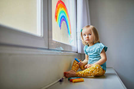 Adorable toddler girl drawing rainbow on window glass as sign of hope. Creative games for kids...