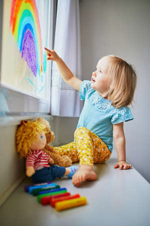Adorable toddler girl drawing rainbow on window glass as sign of hope. Creative games for kids...
