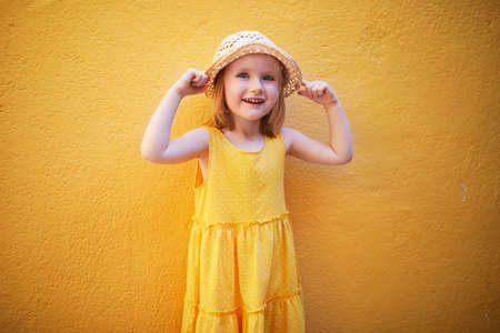 Cheerful three years old girl in yellow dress and straw hat posing against yellow wall. Happy child on a summer day. Summertime activities or vacation conceptの写真素材