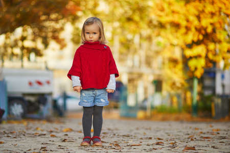 Adorable little girl in red poncho walking in autumn park on a sunny fall day. Happy child having fun outdoors on a street of Paris, France. Outdoor autumn activities for kidsの写真素材