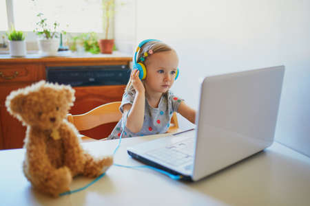 Toddler girl wearing eraphones with laptop. Kid using computer to communicate with friends, elderly relatives or kindergartners. Education or online communication for kids. Stay at home entertainmentの写真素材