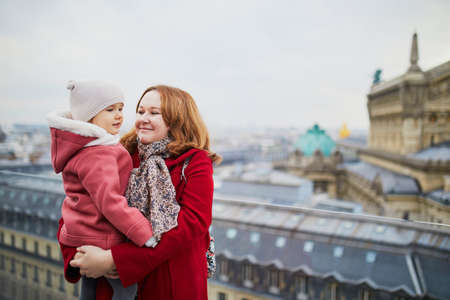 Young woman holding adorable todler girl while enjoying the view from Parisian rooftop terrace. Mothe and daughter in Paris, Franceの写真素材