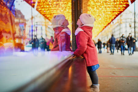 Toddler girl looking at window glass of large department store decorated for Christmas. Child enjoying holiday season in Paris, Franceの写真素材