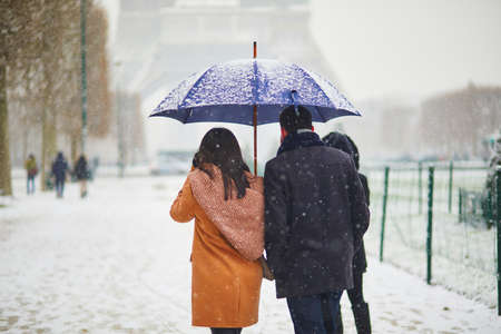 Couple passing by the Eiffel tower in Paris on a day with heavy snow. Unusual weather conditions in Paris, France. Romantic trip to France at winterの写真素材