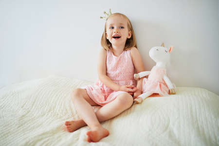 Adorable little girl in pink dress and golden crown dressed as princess playing with unicorn. Kid having fun with soft toy. Child playing with stuffed toyの写真素材