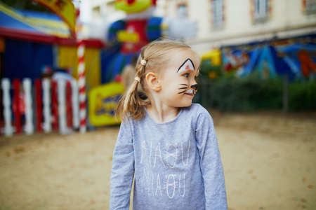 Little preschooler girl with cat face painting, making funny grimace outdoors. Children face...