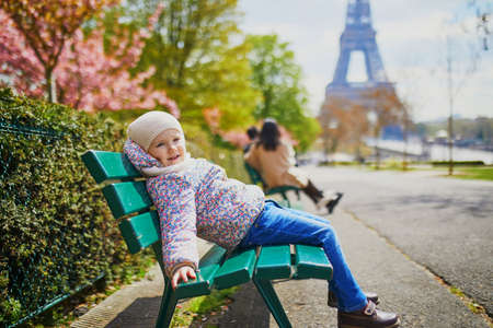 Adorable three year old girl sitting on the bench near the Eiffel tower in Paris, France. Cherry blossom trees in full bloom in Paris on a spring dayの写真素材