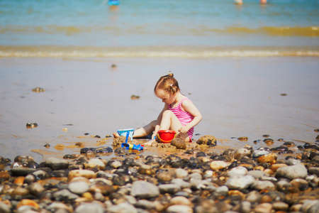 Adorable toddler girl playing on the sand beach at Atlantic coast of Brittany, France. Small child enjoying vacation by the sea or ocean. Travelling with kidsの写真素材