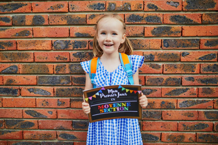 Cheerful 4 years old girl holding a poster with text My first day in kindergarten written in French....