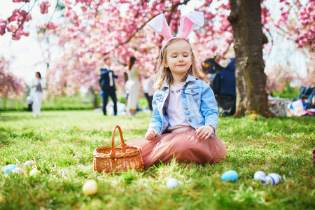 Preschooler girl wearing bunny ears playing egg hunt on Easter. Child gathering colorful eggs in basket. Little kid celebrating Easter outdoorsの写真素材