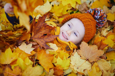 Adorable preschooler girl enjoying nice and sunny autumn day outdoors. Happy child lying on the ground covered with utumn leaves in Paris, France. Outdoor fall activities for kidsの写真素材