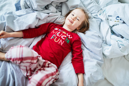 Happy preschooler girl wearing Christmas pajamas, playing on bed. Celebrating seasonal holidays with...