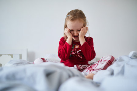 Unhappy preschooler girl wearing Christmas pajamas, sitting on bed. Celebrating seasonal holidays with kids at homeの写真素材