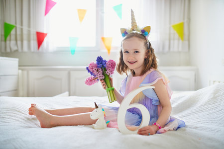 Adorable preschooler girl in red dress sitting on bed with pink hyacinths and white wooden number six. Sixth birthday concept. Kid celebrating sixth birthdayの写真素材