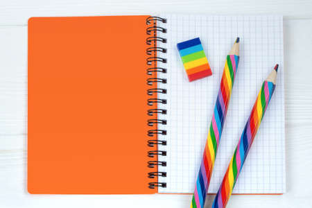 Open copybook with two rainbow-coloured pencils and a rubber on white wooden table