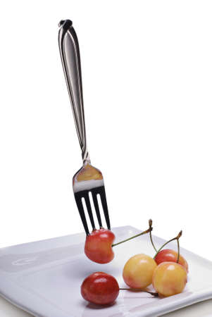 A fork pierces a floating Rainier cherry.  Other Ranier cherries rest on a white plate against a white background.の写真素材