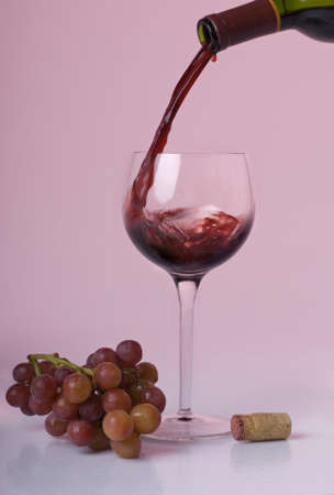 Red wine pouring from a bottle into a wine glass with red grapes and a cork beside the glass on a rose colored background.の写真素材