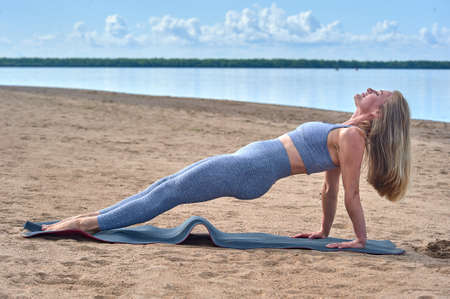 A woman does yoga on a sandy beach against the background of a river