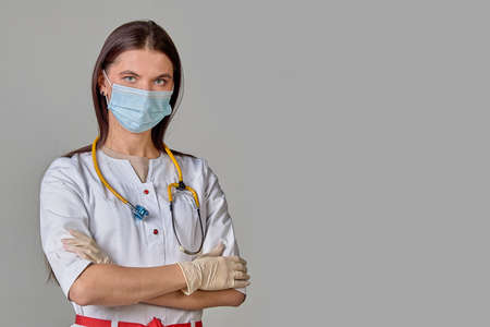 A young female doctor in a white coat with a stethoscope and a medical mask on a gray isolated backgroundの写真素材