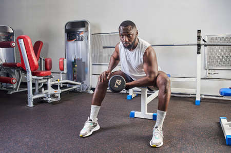 An African American man shakes his biceps while sitting on a bench in the gymの写真素材