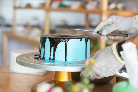hand of pastry chef pouring melted chocolate at blue berry cake on the wooden table inside bakery...