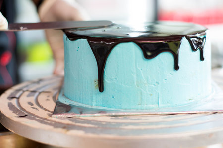 zoom of pastry chef hand using rubber gloves smooth over melted chocolate on round blue cake with...