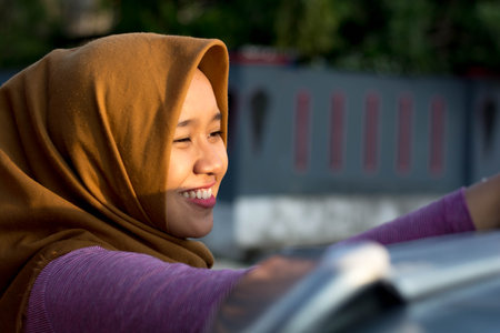 close up shot portrait of a hijab woman washing car roof and smilling in front of her house on a sunny dayの写真素材