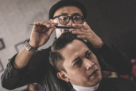 close up portrait of barber hairstyle styling hair of his costumer hair who sitting in chair with...