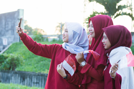 portrait of young asian muslim woman group with indonesian flag taking group selfie and celebrate...