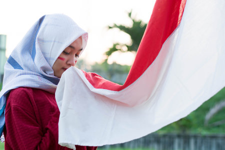 portrait of young asian muslim woman with scarf kissing red and white flag of indonesia to celebrate...