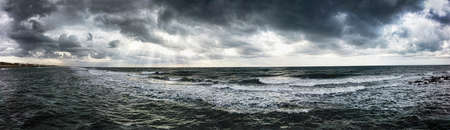 Dramatic weather panorama over the sea threatening waves crashing at the shore with overcast sky and sunbeams on the waterの写真素材
