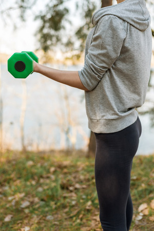 Girl in sports pants and hoodie standing up shakes her bicep hands with dumbbellの写真素材
