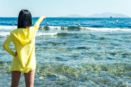 Travel and vacation, a girl with black hair in a yellow jacket stands against the blue sea and shows her hand towards the horizon. Horizontal frameの写真素材