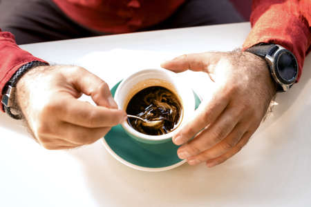 Man holding a pink ceramic cup of hot black coffee in his hands, sitting at the wooden table in a...
