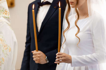 The bride and groom hold shining candles during the ceremony in the church. Hands of newlyweds with candles in the church. Church religious detailsの写真素材