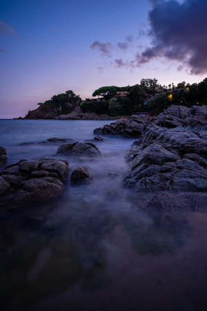 rocks on beach with small town in the background at dusk and silk effect, blanes catalonia spain