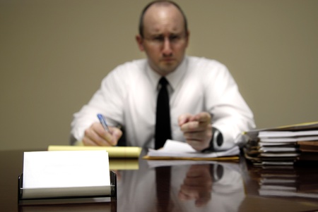 Stern Man sitting at desk holding pen and pointing finger at viewer with blank business cardの写真素材