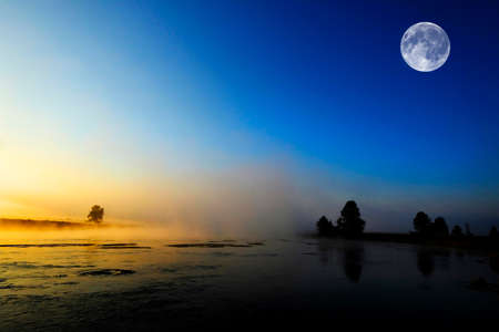 Morning sunrise with trees and river under blue sky and glowing mist and full moonの写真素材