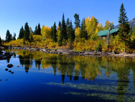 Old cabin in forest of trees in Autumn fall by river water reflectionsの写真素材