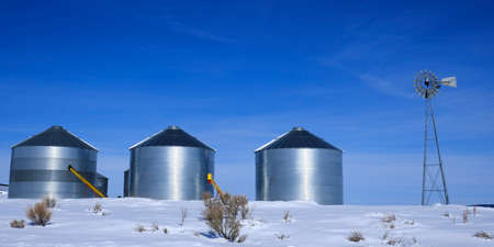 Windmill and steel grain silos in winter snow with blue sky on farm for agricultural farmingの写真素材