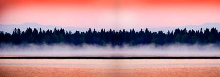 Sunrise or sunset on a lake with mist rising from the water and pine trees mountains in background...