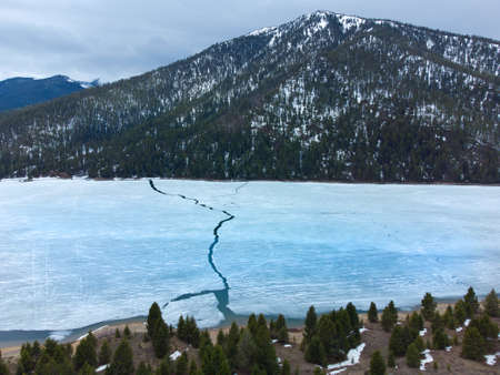 Aerial view from a drone flying above a frozen lake water with cracked ice and mountains forestの写真素材